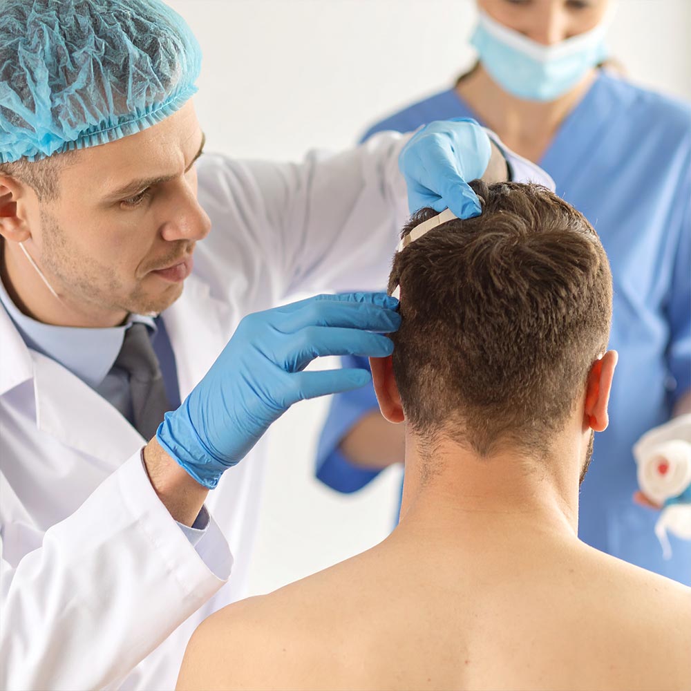 Ein Arzt legt einem sitzenden Mann einen Verband am Hinterkopf an, unterstützt von einer Pflegekraft im Hintergrund.