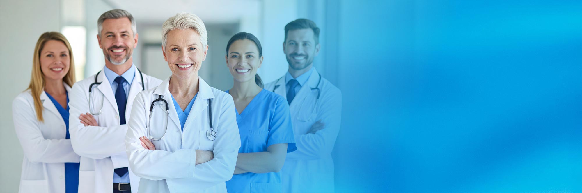 Ärztliches und medizinisches Fachpersonal steht lächelnd in einer Klinik oder Praxis, im Vordergrund eine Ärztin mit verschränkten Armen, dahinter ein Team aus Ärzten und Pflegekräften vor hellem, modernem Hintergrund.